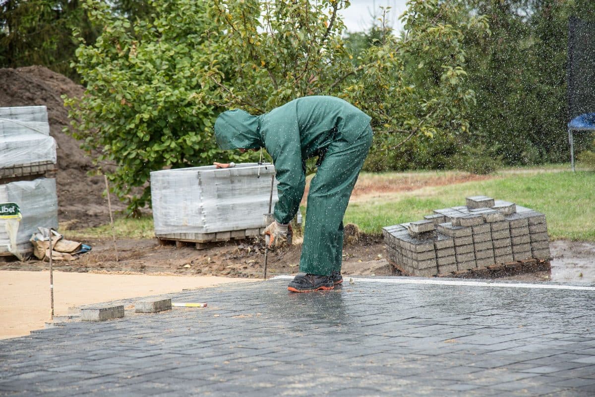 Arbeiter in Regenkleidung verlegt Pflastersteine, dahinter Paletten und Garten